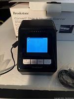 Front view of the Brookstone iConvert Slide and Negative Scanner powered on showing a blue preview screen. Control buttons visible at the front, placed in front of its original box.