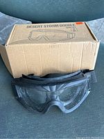 Photo showing black Desert Storm goggles in front of a cardboard box labeled 'Desert Storm Goggle Lens Set'. Goggles show some dust and dirt, need cleaning.
