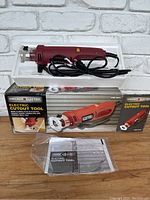 Electric cutout tool, packaging box, and owner's manual on wooden surface with white brick background.