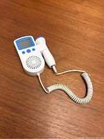 White and blue fetal doppler device lying on wooden surface, showing LCD screen, buttons, speaker and connected probe by coiled cord.