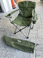Green fold-up camping chair opened and set up on stone floor with visible cup holder on right armrest and storage pouch on left armrest, shown with matching green ULINE carry bag beside it.