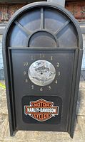 Full front view of black Harley-Davidson wall clock showing arched top, clock face with gold numbers, silver emblem with motto, and Harley-Davidson logo panel below.