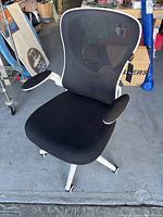 Front view of Hbada office chair showing black mesh backrest, padded black seat, white base with armrests, on concrete floor with some background boxes.