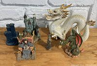 Photo showing a white dragon figure, a green dragon, castle models, and a figure on a throne on a wooden surface against a white brick wall.