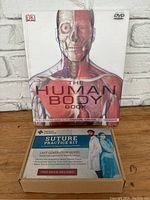 Front view of The Human Body Book with anatomy illustration and DVD logo. Next to it is the box for the surgical suture practice kit with promotional text.