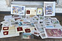 Wide view of various postal stamps and first day covers laid out on a surface, includes framed collectible stamps and loose sheets.