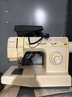 Front view of cream-colored Singer sewing machine with foot pedal and stitch pattern options visible.