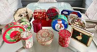 Front perspective of all 14 assorted Christmas tins on a glass table showing various shapes, holiday-themed prints, and colors.