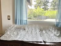 Wide view of assorted crystal stemware and glassware arranged on table near window, showing many glasses of various styles and sizes.