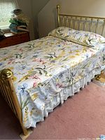Comforter set with white background featuring large floral pattern in yellow, blue, pink and green colors, laid on brass bed frame.