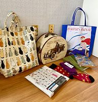 Displays all lot items, including the vintage automobile box, cat tote, Harper's Bazaar tote, shower curtain packaging, socks, and journals arranged on wooden surface.