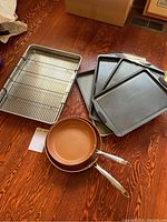 Five nonstick baking sheets stacked, two steel nonstick frying pans with copper-colored interior, and a large roasting pan with wire rack inside on wood floor.