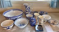 Assorted blue and white dishware laid out on a table including bowls, mugs, a jug, vases, and plates with floral and geometric designs.