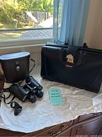 Photo showing the briefcase and Bell & Howell binoculars with their leather case placed on a table near a window.