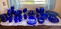 Wide view of all cobalt blue glassware arranged on a wooden surface under a window with lace curtains. Items include bowls, plates, mugs, vases, goblets, serving dishes, and a water jug.