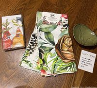 Photo showing two botanical print cotton tea towels, package of cardinal bird printed napkins, and green glazed pottery dish on wooden surface.