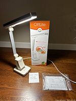 Full view of the white OttLite Slimline task lamp plugged in, positioned on wooden floor. Lamp is lit showing slim light bar. Original box in background, plastic sleeve with instructions in foreground.