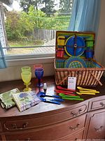 Photo showing the full picnic set including basket, plates, colorful tumblers, utensils, cloth, and napkins on a wooden surface near window.