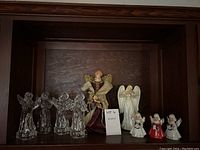 Shelf display of 10 angel figurines including 4 clear glass angels, large burgundy and gold angel, white angels, and small colored angels.