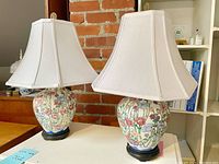 Pair of matching ceramic floral Japanese lamps with white fabric shades shown on white table with brick background.