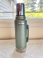 Light green Stanley Aladdin thermos with textured finish and metal cup lid, standing upright with handle visible on the side.