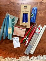 Photo showing an assortment of boxed and loose tapered candles in various colors including blue, pale blue, red, green, white, and patterned candles on a wooden floor.
