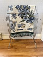 Front view of silver 4-tier wire towel and blanket stand holding a woven blanket with floral pattern.