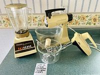 Overview photo showing the Osterizer blender, Sunbeam Mixmaster stand mixer with bowls and attachments, and handheld mixer together on a countertop.