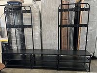 Wide shot of two black metal coat racks side by side, showing full height, multiple hooks on vertical backing bars, and shoe storage shelves below bench surface.