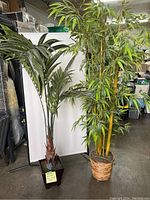 Two artificial plants side-by-side; faux palm tree with wide green fronds and bamboo plant with many tall green stalks and leaves