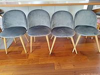 Front view of four grey velvet upholstered dining chairs with slender wood-look metal legs on wooden floor.