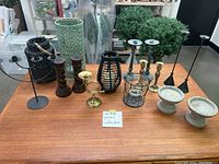 Photo showing 16 assorted lanterns and candle holders arranged on a wooden table including black metal lanterns, green ceramic lantern, spiral carved wooden candle holders, brass holders, and ceramic pieces.