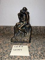 Front view of bronze-coloured sculpture depicting two nude lovers embracing, seated on textured base.
