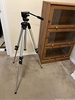 Full view of the tripod showing silver aluminum legs with black leg locks and the mounting head, set up on carpet indoors near a wooden cabinet.