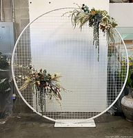 Full front view of large white round metal grid wedding arch with some floral arrangements attached (not included). The arch has a flat base and is standing in a warehouse.