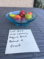 Aqua blue glass bowl containing assorted artificial fruit including a banana, apples, pears, cherries, blackberries, and oranges, photographed outside on brown surface with label showing lot number and description.
