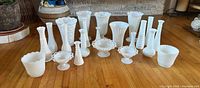 Photo of 22 assorted milk glass vases and decorative bowls arranged on wooden floor. Pieces vary in size, shape, and pattern.