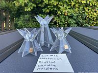 Three clear starburst crystal candle holders arranged on outdoor surface with greenery background. Two smaller holders have Vintage Nachtmann stickers. Handwritten note visible. Water droplets on surface from rinsing dust.