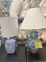 Two oriental table lamps side by side on a black surface, one with a square shaped base and cylindrical shade, the other double-gourd shaped base with wooden stand and white conical shade.