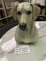 Front view of the yellow Labrador retriever sculpture showing detailed facial features and the dog's head shape, placed on a marble surface with a note indicating the artist and dog breed.