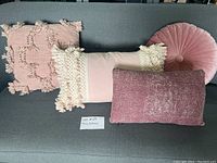 Four pink and purple pillows arranged on a gray sofa including a round velvet pillow, a rectangular crushed velvet pillow, a rectangular pillow with cream tassels, and a square pillow with tassels and knotted loops.