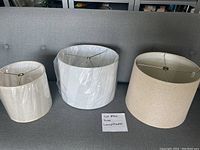 Three lampshades displayed on a grey sofa: small cream, large cream, and stark white. Two shades are wrapped in plastic.