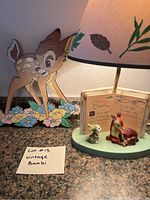 Full view of Bambi lamp with ceramic figurines in front of a book-themed backing and original leaf-patterned lampshade, next to the large colorful Bambi cutout on floral ground.