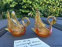 Two amber blown glass swan candle holders side by side on a dark table with greenery background, showing front and side angles.