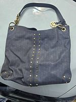 Front view of a grayish-blue Michael Kors Astor leather shoulder bag with signature MK monogram pattern and gold-tone studs along the front and corners. Leather and gold chain strap visible with MK logo charm.