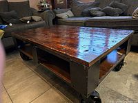Angled side view of the Pinebrook coffee table showing polished wooden top, black metal frame, and large caster wheels.
