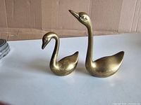 Two vintage brass swan figurines displayed on a white surface with a plain backdrop. One swan is larger with an upright neck, the smaller swan has a lower neck position.