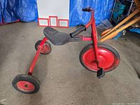Side angle of vintage heavy tricycle showing red metal frame, large front wheel with pedals, and body seat.