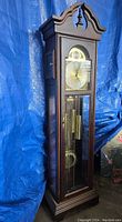 Full vertical view of the grandfather clock showing wooden case, glass front and clock face with arched top and finial.