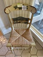 Front view of the wooden chair with cream paint, showing the woven wicker seat and the hand painted scene on the backrest with horse-drawn sleigh and houses in winter.